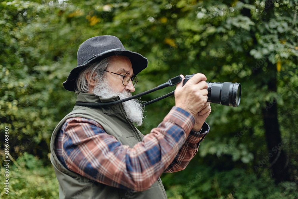 Fototapeta premium Caucasian senior man holding digital camera while photographing nature in forest. Image suitable for active retirement concepts, travel marketing, outdoor hobby advertising, side view