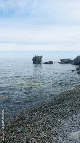 Rocky shoreline of Lake Baikal with distinctive rock formations rising from calm clear water, pebble beach and serene natural landscape under cloudy sky