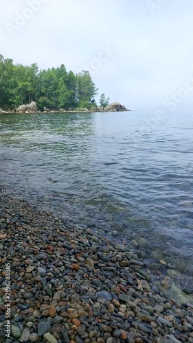 Rocky shoreline of Lake Baikal with distinctive rock formations rising from calm clear water, pebble beach and serene natural landscape under cloudy sky