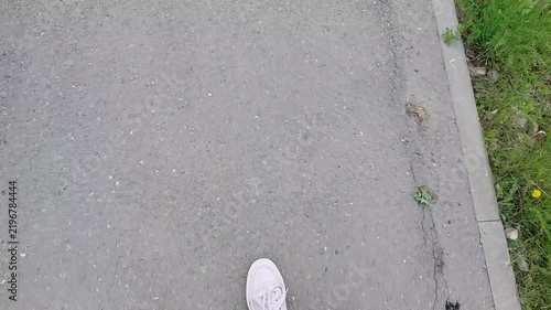 First-person view of feet in white sneakers and blue jeans walking along an asphalt path bordered by green grass and dandelions
