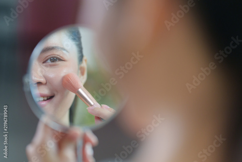 Asian woman applying blush with makeup brush in mirror for cosmetics beauty routine