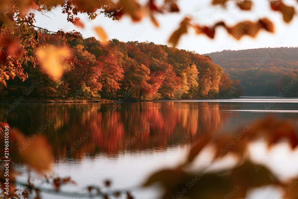 Obraz premium Autumn landscape with colorful foliage, trees, and lake reflection viewed through frame of leaves