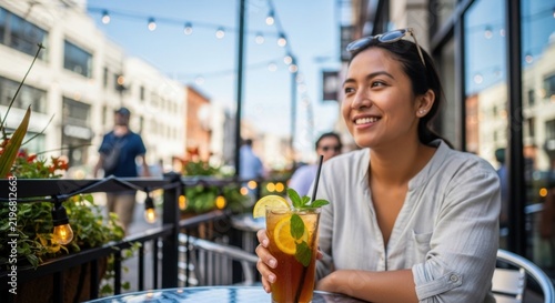 Smiling young woman enjoying a refreshing iced tea drink on a sunny outdoor terrace in the city