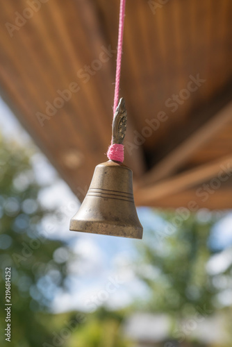 Obraz na plátně Close-up of a small brass bell hanging from a colorful cord beneath a wooden structure outdoors