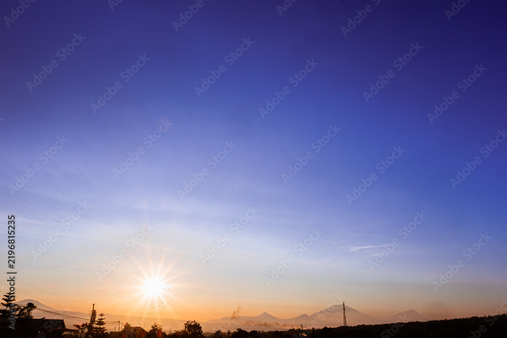 Obraz premium A stunning sunrise landscape with a bright sunburst effect on the eastern horizon, showing the silhouette of the countryside, trees