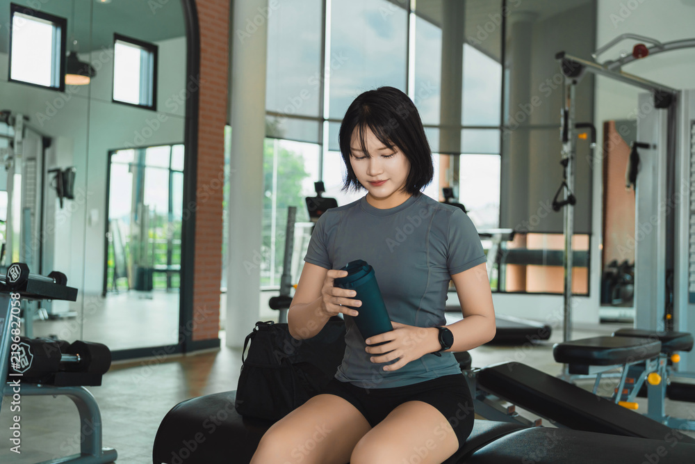 Obraz premium Young woman sitting on bench in gym holding water bottle resting after workout routine in modern fitness center