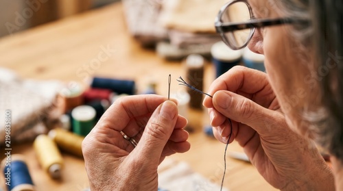 presbyopia woman stress fatigue concept. Elderly hands threading a needle with colorful sewing supplies nearby.