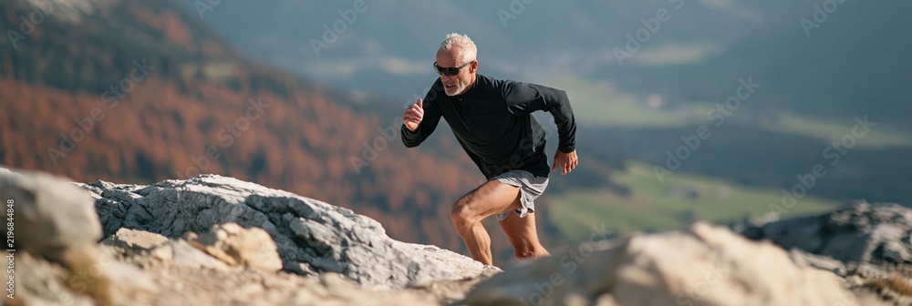 Obraz premium Active mature man trail running along rocky mountain ridge in autumn for outdoor fitness banner and landscape background