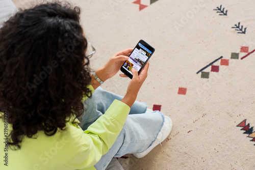 Young African-American woman browsing travel blog on mobile phone at home, to...