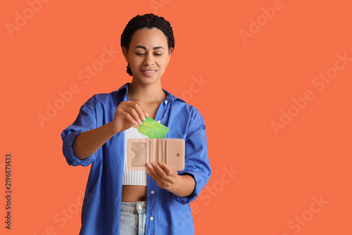 Young African-American woman putting credit card into wallet on orange backgr...