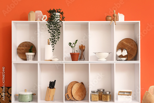 Shelf unit with kitchen utensils and plants near orange wall