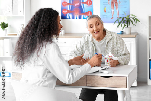 Female doctor with pill bottle showing spine model to senior patient at table...