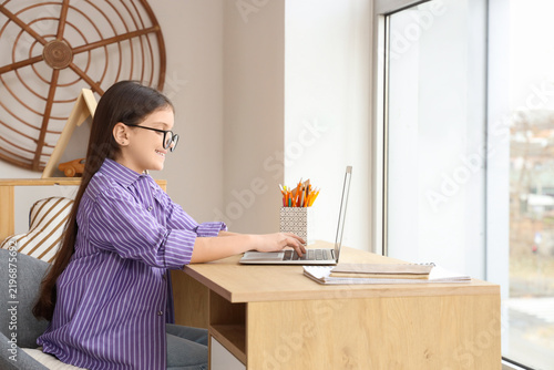 Teenage girl in eyeglasses using laptop on table at home
