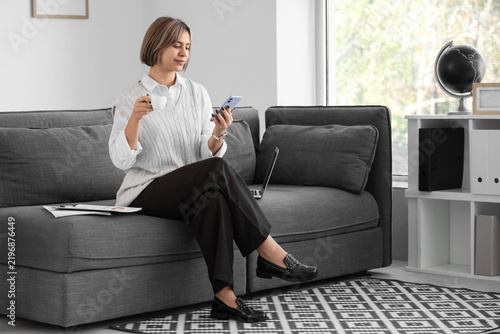Successful businesswoman with cup of coffee using mobile phone in office