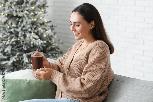 Happy young woman with glass cup of hot cocoa sitting on sofa in living room ...