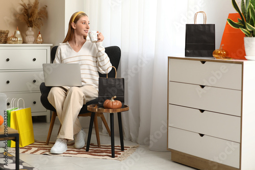 Young woman with laptop and tea cup shopping online in armchair at home