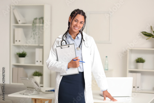Young African-American female doctor with clipboard near table in clinic