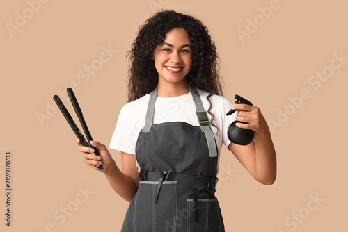 Beautiful young happy African-American hairdresser with hair straightener and...