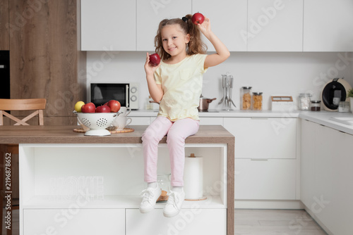 Cute little girl with red apples sitting on table in kitchen