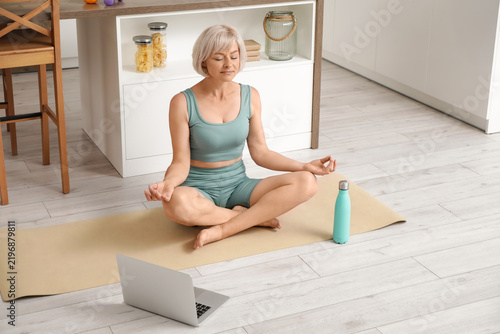 Sporty beautiful mature woman with laptop and bottle of water meditating in k...