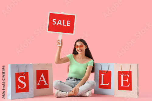 Beautiful young happy woman with shopping bags and SALE sign sitting on pink ...