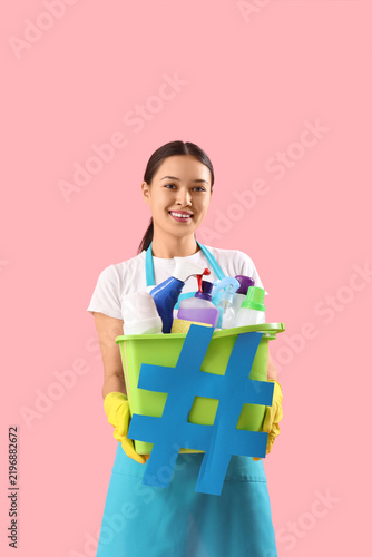 Female janitor with cleaning supplies and hashtag sign on color background