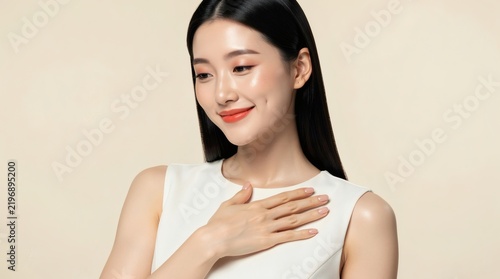 Woman with clean skin smiles gently while placing her hand on her chest against a neutral beige background.