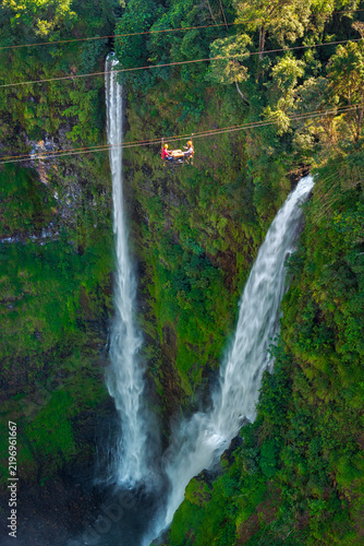 Foto Aerial view of Tad Fane Waterfall in Laos, with a tourist enjoying tea while suspended on a zipline above the spectacular twin cascades and lush rainforest of the Bolaven Plateau