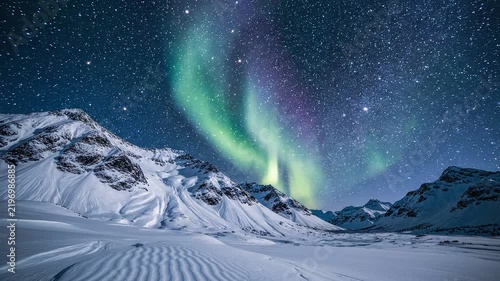 Vibrant green and purple aurora borealis lights up the night sky above a snow-covered mountain range under a starry sky.