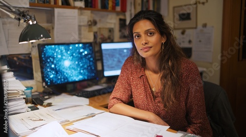 Focused female student working late at her desk in a cozy study environment, surrounded by papers and computer screens displaying vibrant cosmic images, reflecting a studious yet calm atmosphere.