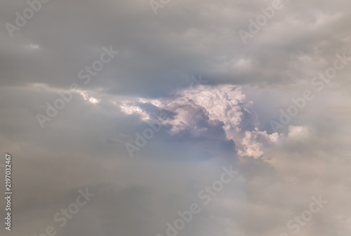 A striking dramatic sky featuring a dense layer of dark grey storm clouds breaking open to reveal textured, brightly illuminated cumulus formations behind. Soft natural sunlight highlights the billow