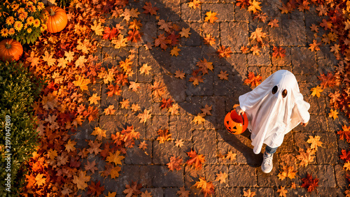 An overhead view shows a child in a white sheet ghost costume walking along a leaf-covered path with an orange pumpkin bucket, surrounded by scattered autumn foliage and seasonal decorations outdoors