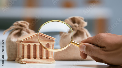 A hand with a magnifying glass examines a wooden model of a house next to bags of money, symbolizing real estate analysis and mortgage investment.