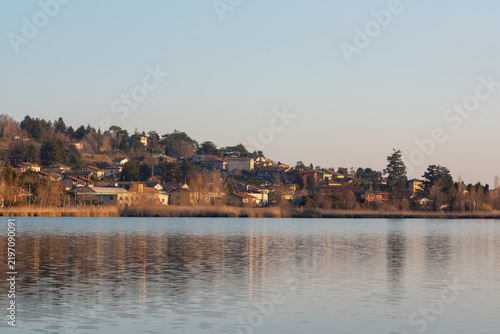 A serene landscape photograph captures a tranquil lakeside village nestled on a gentle slope under a clear, pale blue sky during the golden hour