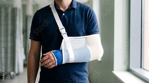 Man with broken arm in blue plaster cast and white mesh medical sling standing in a clinic for healthcare recovery and personal injury insurance claim concept.