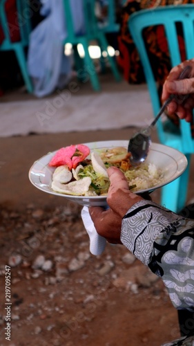 Canvas Print Guest wearing a traditional patterned batik shirt holding a plate of Indonesian festive food with crackers and rice at a social gathering