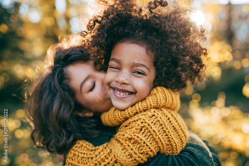 Joyful mother hugging her happy little daughter outdoors in a park during autumn sunlight