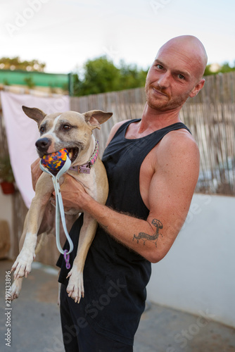 Konstfotografi Man with a tattoo on arm holding a domestic dog with a colorful toy in its mouth