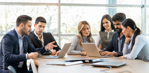 Business executives sitting around conference table in office explaining corporate project details on laptops.