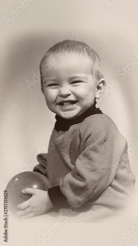 A little boy in a vintage photograph is holding a ball, posing for the camera, smiling, and laughing.