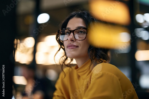 Thoughtful young woman wearing glasses and yellow top sitting indoors