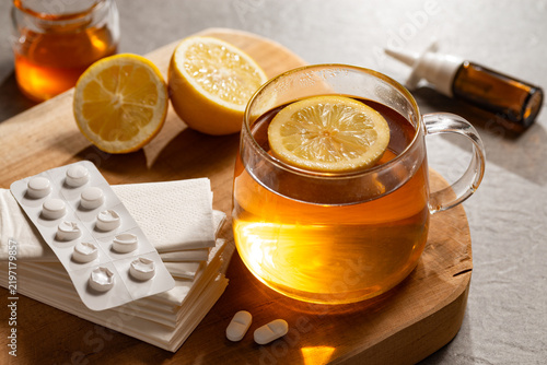 Erkältungszeit - Heißer Tee in Glastasse auf Holzbrett mit Zitronen, Nasenspray und Tabletten
