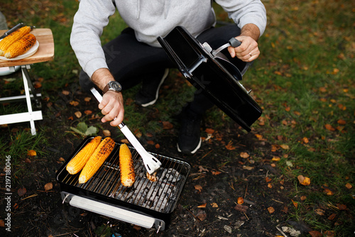 Фотография Cropped closeup of man cooking sweet corn cobs on portable charcoal grill with tongs at campsite in autumn forest, holding barbecue lid