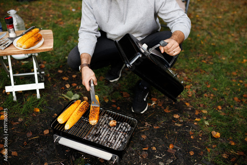 Картината върху платно Cropped closeup of man cooking sweet corn cobs on portable charcoal grill with tongs at campsite in autumn forest, holding barbecue lid