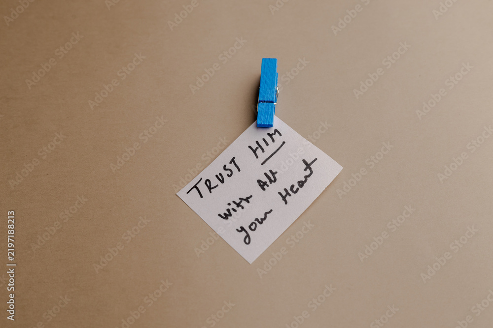 Obraz premium A small white note with handwritten Christian quote attached by a blue clothespin to a neutral background, inspiring trust and faith message