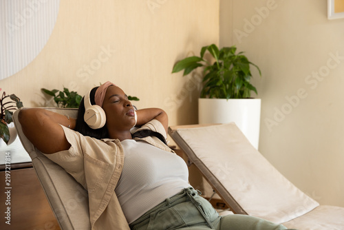 African adult woman reclining on padded lounge chair in relaxation nook wearing over-ear headphones