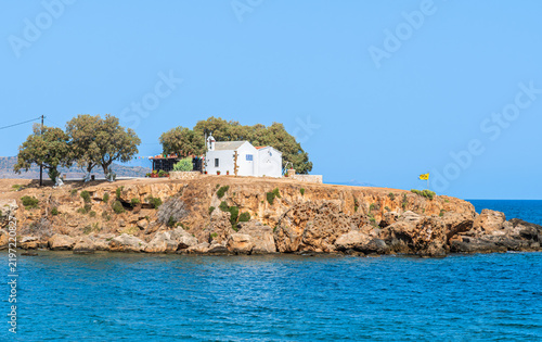 The Chapel of the Holy Apostles at Agii Apostoli beach in Chania, Crete, a historic white orthodox church on a rocky peninsula in summer.