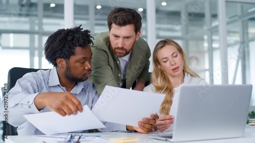 Three multiracial business partners concentrated formal team managing papers documents with laptop corporate service in office colleagues multiethnic group working paperwork analysing using computer