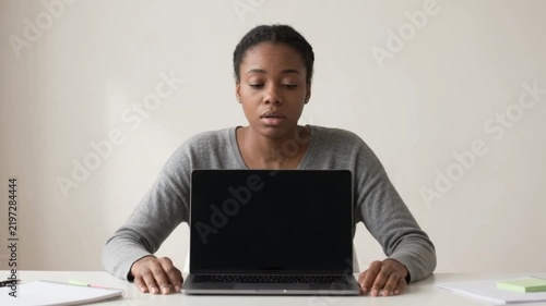 A young Black woman sits at a desk with a laptop in front of her, looking down with a thoughtful expression.