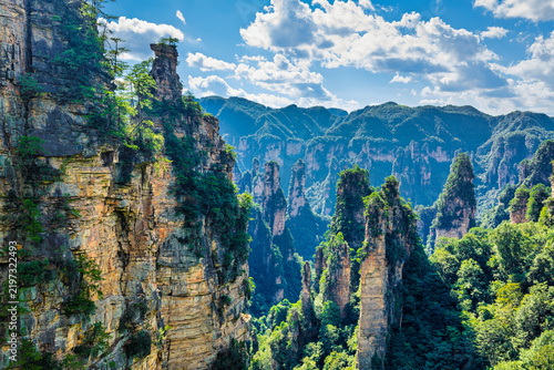 Tall sandstone pillar peaks rise above lush green forested valleys under bright blue sky with cumulus clouds in Zhangjiajie landscape.